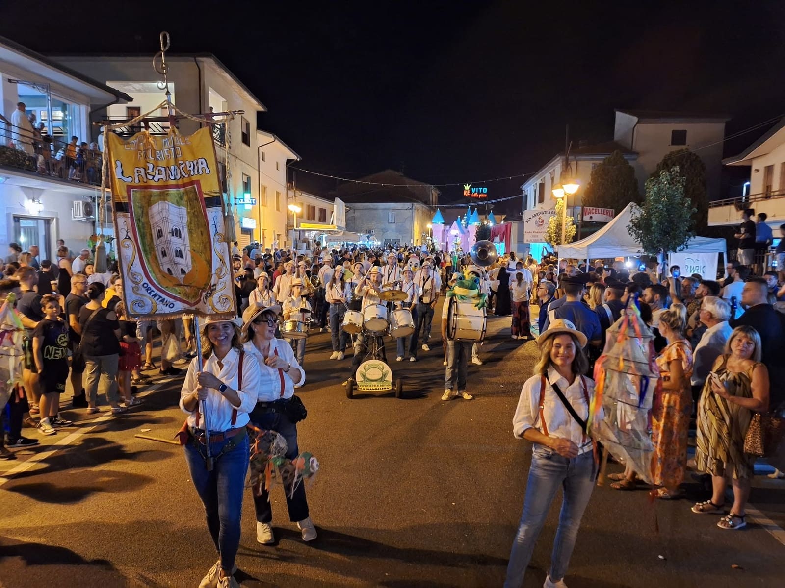 Die Folklore-Kapelle La Ranocchia zieht an einem Sommerabend durch Orentano, Banner erhoben, Trommeln und Frosch-Maskottchen in der Menge